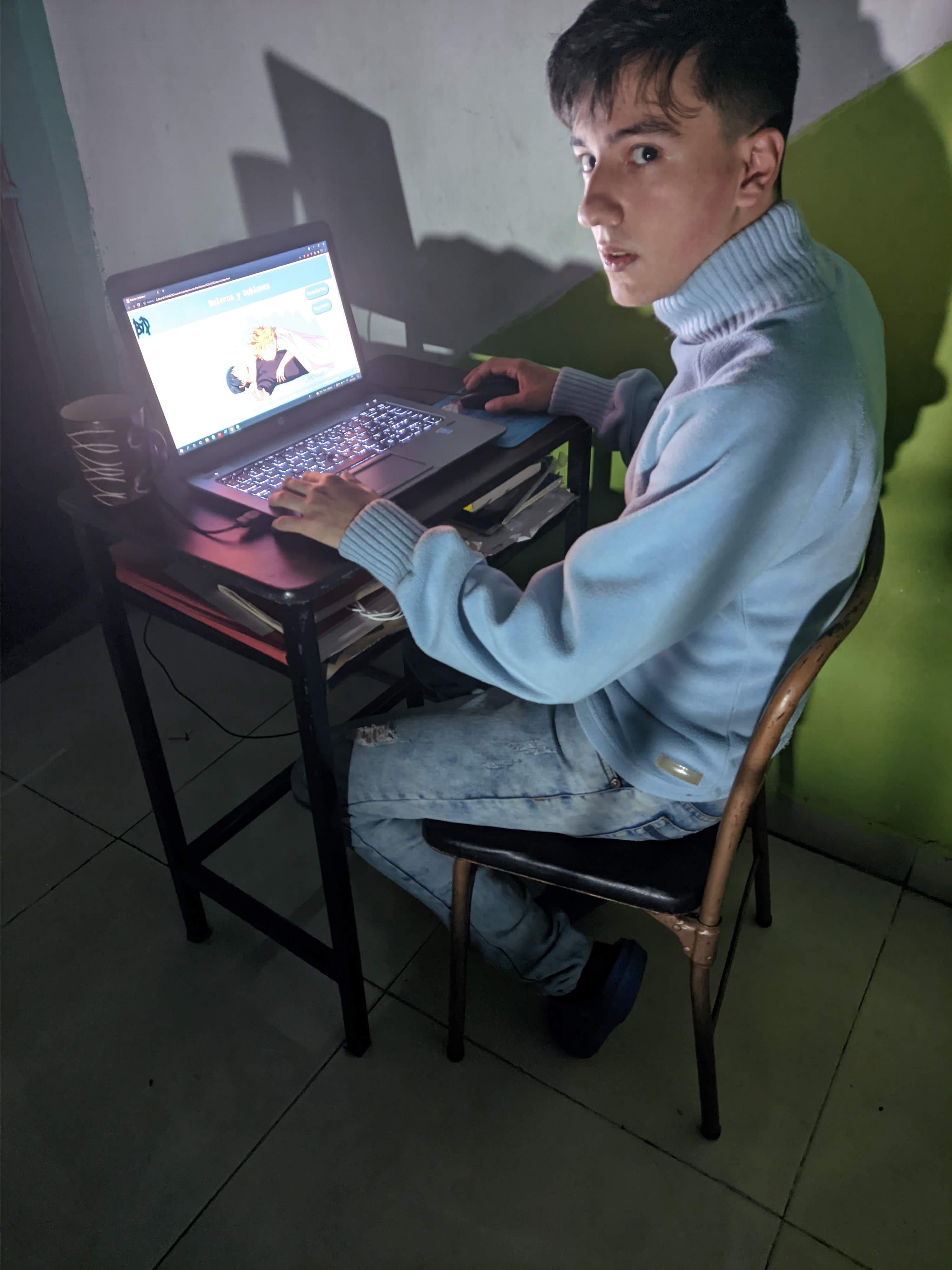 Yo de noche en un escritorio pequeño contra una pared verde, volteando a la cámara, con la página terminada abierta en la laptop.
