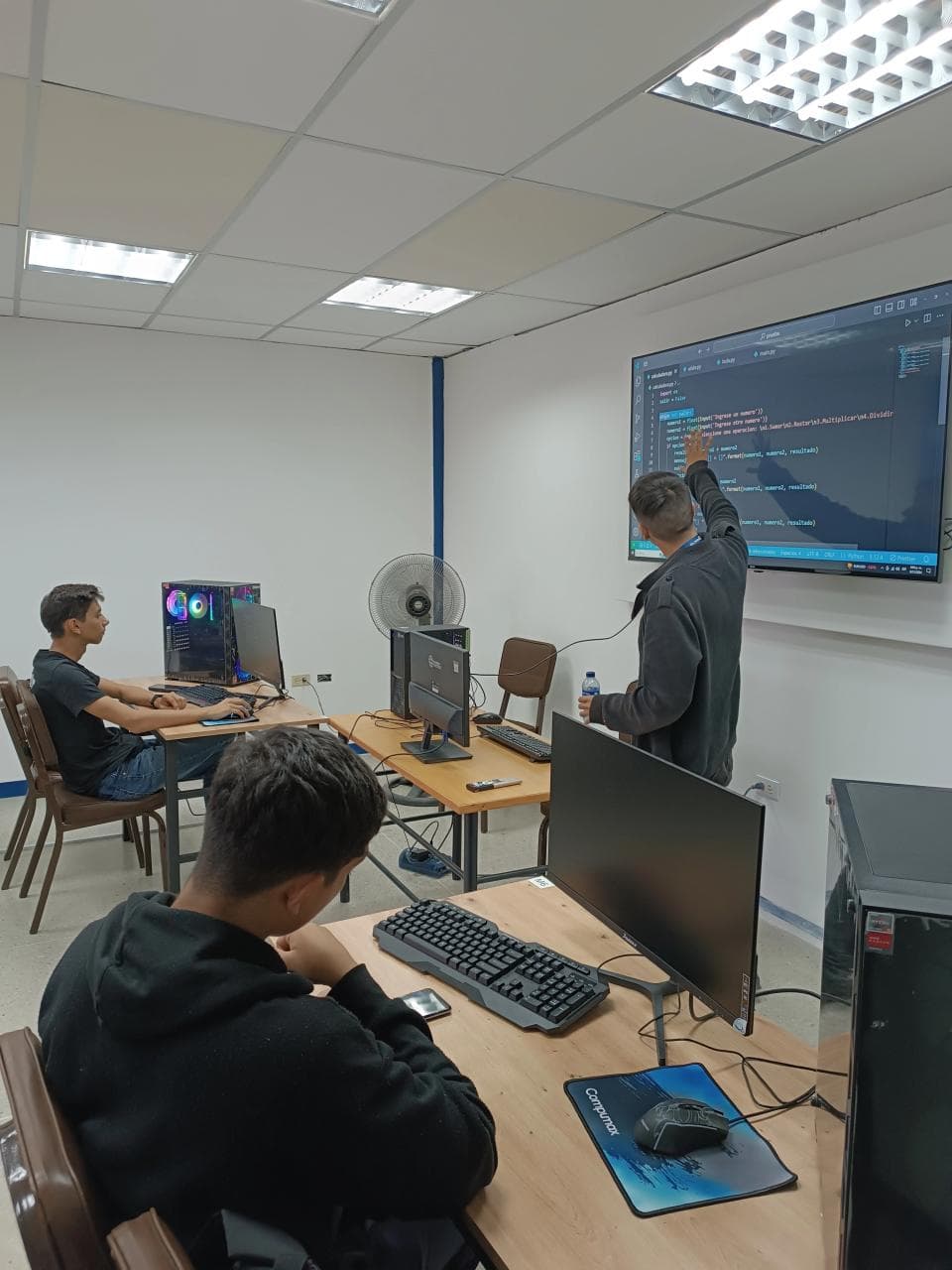 Un laboratorio de computación: estoy frente a la pantalla grande explicando un ejercicio de Python mientras dos estudiantes siguen la clase desde sus puestos.