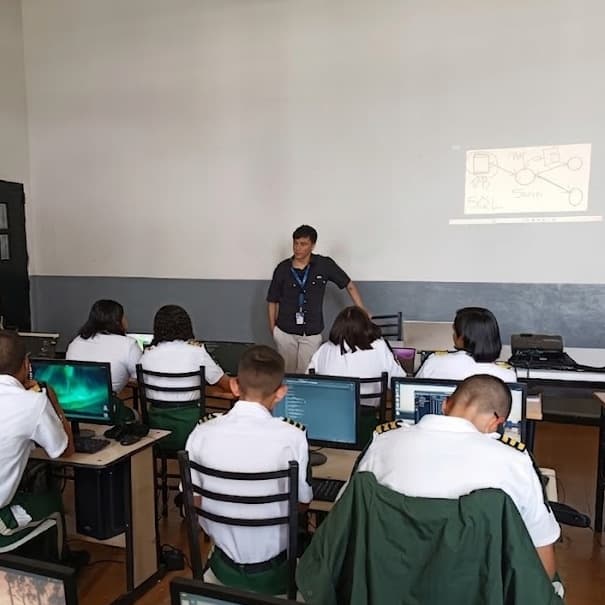 Un aula con estudiantes uniformados frente a computadoras, y yo adelante junto a un diagrama proyectado en la pared.