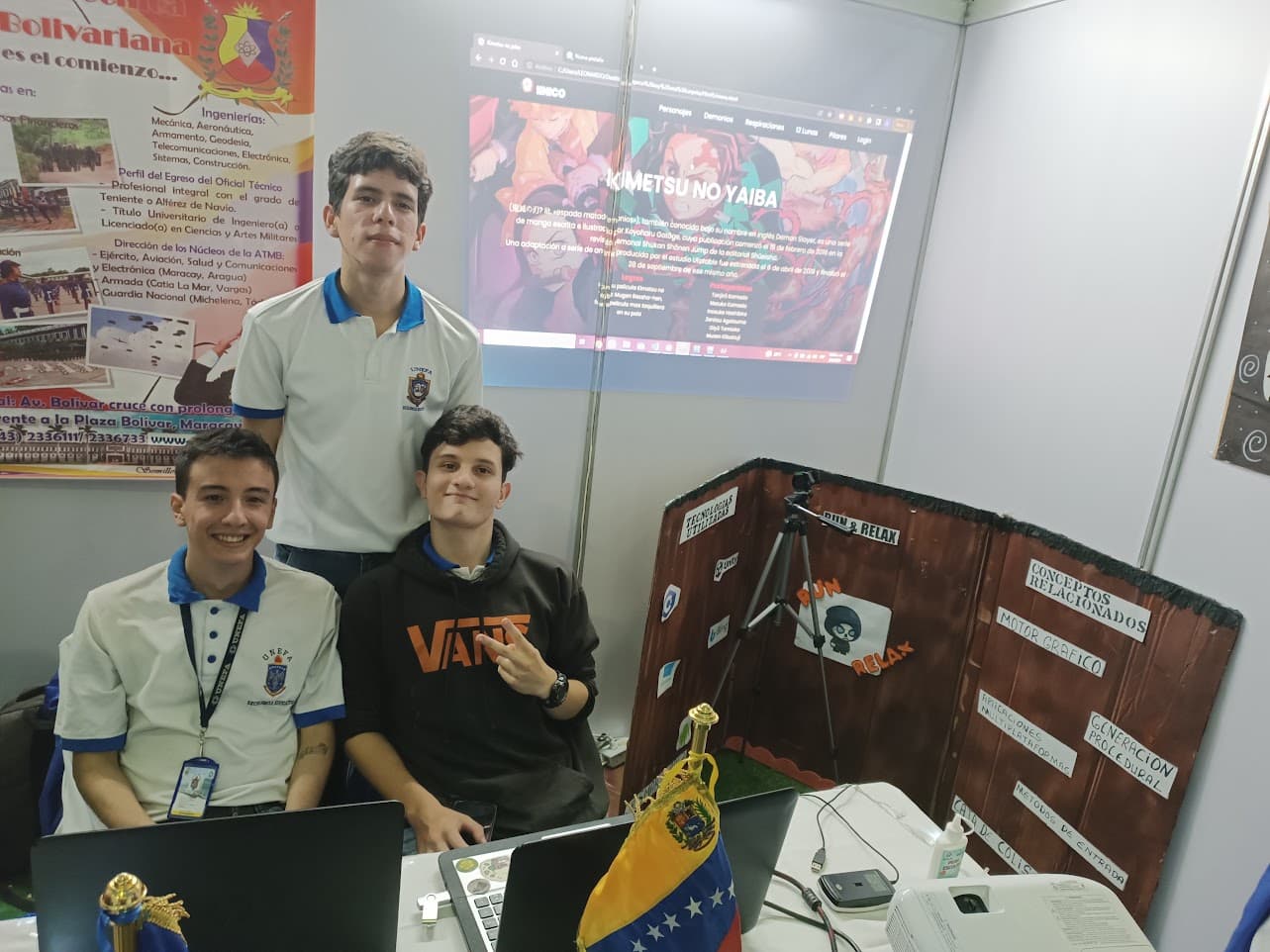 Tres de nosotros en un stand de una feria universitaria, con nuestro proyecto proyectado detrás y un cartel hecho a mano que lista las tecnologías que usamos.